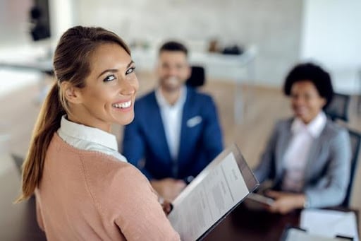 Imagem de uma profissional sorrindo com uma pasta na mão, em uma sala com outros profissionais para simbolizar o que é departamento pessoal